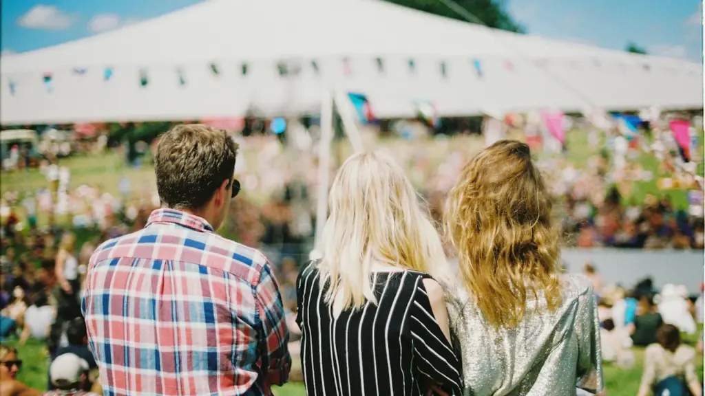 Trīs jaunieši no aizmugures skatās uz brīvdabas festivāla norisi saulainā vasaras dienā.