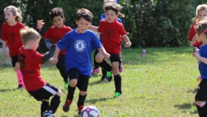 Bērni spēlē futbolu zālienā, attīstot savas sportiskās prasmes un komandas sadarbību.