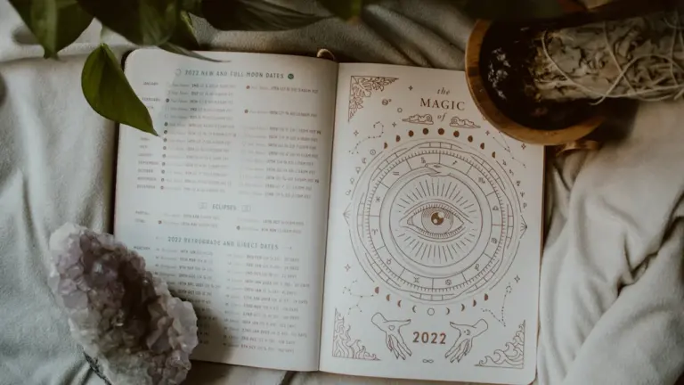 Atvērta dienasgrāmata ar astroloģiskiem simboliem un kristālu uz gultas pārklāja