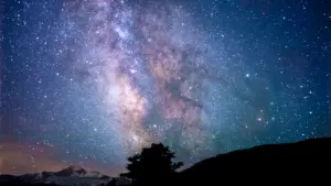 Tumšas debesis ar zvaigžņu segu virs mierīgas kalnu un koku silueta ainavas.