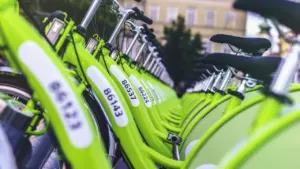 Vairāki zaļi nomas velosipēdi novietoti rindā pilsētas ielas malā dienas laikā.
