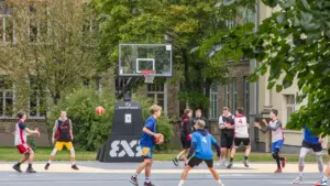 Vairāki jaunieši spēlē basketbolu uz āra laukuma pie skolas ēkas rudens pēcpusdienā.